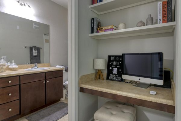 Integra Woods Small desk nook with computer, lamp, and shelves; bathroom with double sinks and towels is visible through open doorway.