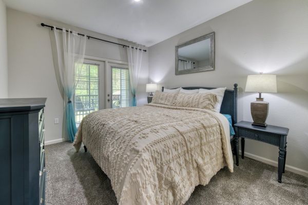 Integra Woods Cozy bedroom with a large bed, two nightstands, lamps, dresser, mirror, and French doors with white curtains leading to a balcony.