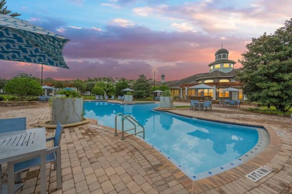 Integra Woods Outdoor pool with patio tables, umbrellas, and a building in the background at sunset with colorful sky.