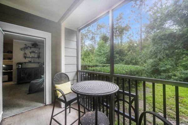 Integra Woods Cozy screened balcony with a round table, chairs, and a view of green trees, next to an open door to a living room.