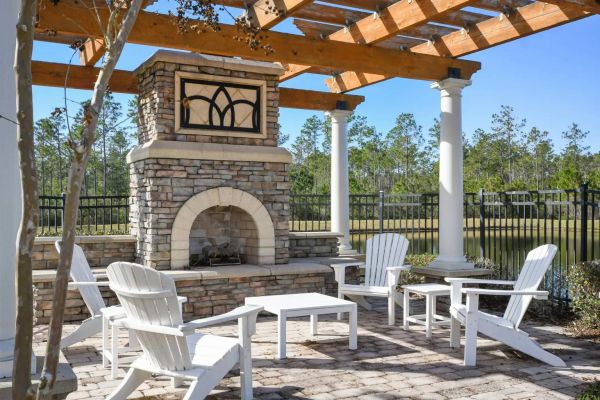 Integra Woods Outdoor patio with white chairs around a stone fireplace under a wooden pergola, trees in the background.