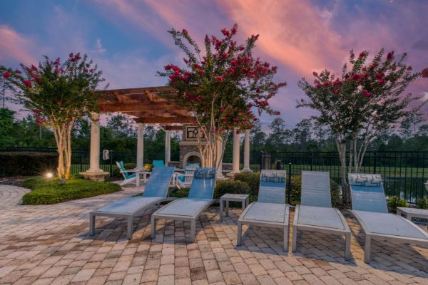 Integra Woods Four poolside lounge chairs, flowering trees, and a pergola with seating under a colorful sunset sky.