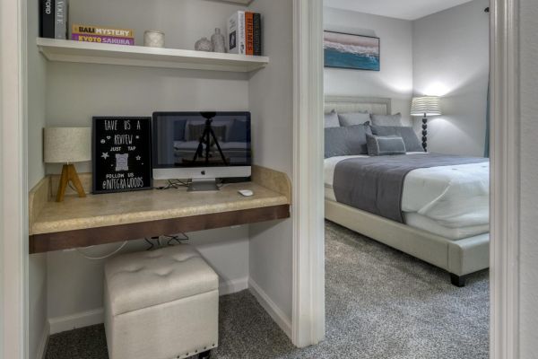 Integra Woods Small home office nook with shelves, a computer, and an ottoman, adjacent to a modern bedroom with a bed and lamp.