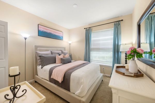 Integra Woods Bright bedroom with a neatly made bed, pastel decor, large window with blue curtains, and a dresser with flowers.